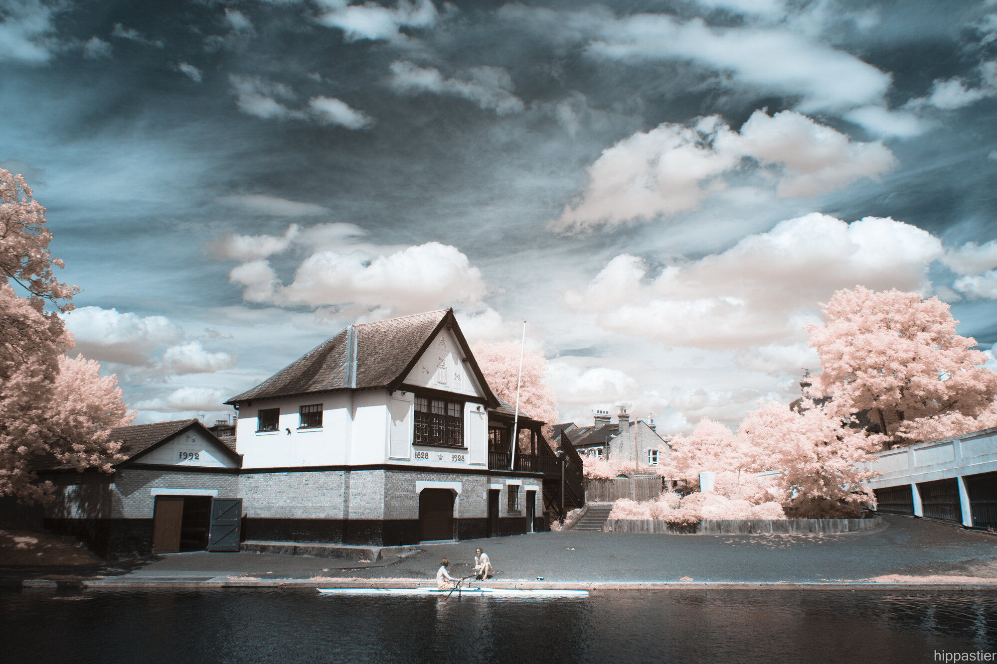 Rowers by a riverside boathouse, pink infrared trees and dramatic sky