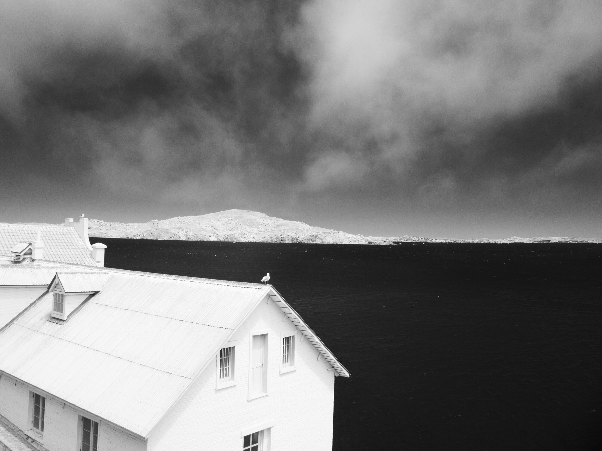 White gabled house on Alcatraz Island against dark water and pale hill, black and white infrared